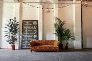 Low sofa next to potted plants and decorative sign with bus frontage in front of white-painted brick wall at 100 Barrington.