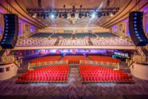 Stage perspective of two-tiered audience seating in Troxy auditorium, with red stalls seats and purple circle seats.