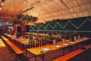 Long wooden tables and benches with leafy decoration laid for seated dinner in cosy space at London Fields Brewery.